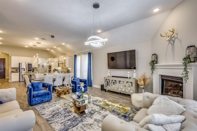 living room with a chandelier, light wood-type flooring, a glass covered fireplace, and vaulted ceiling