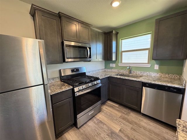 kitchen with dark brown cabinetry, appliances with stainless steel finishes, light stone counters, light wood-style floors, and a sink