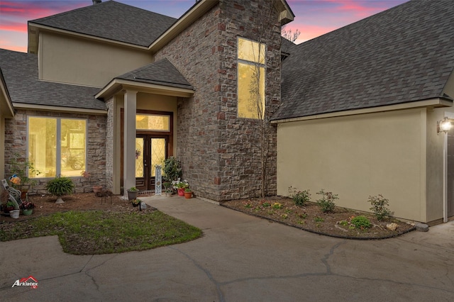 exterior entry at dusk featuring stone siding, roof with shingles, stucco siding, and french doors