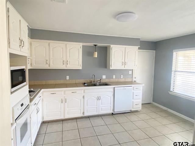 kitchen with a sink, white appliances, light tile patterned floors, baseboards, and white cabinetry