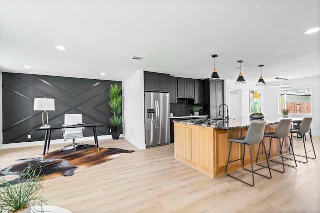 kitchen featuring visible vents, light wood-style floors, a kitchen island with sink, dark stone counters, and stainless steel fridge with ice dispenser