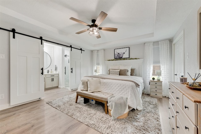 bedroom featuring a tray ceiling, ensuite bath, light wood finished floors, and a barn door