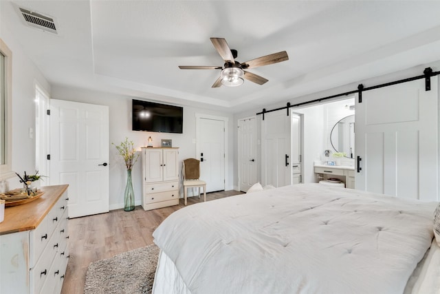 bedroom with a tray ceiling, visible vents, light wood-style flooring, a barn door, and a ceiling fan