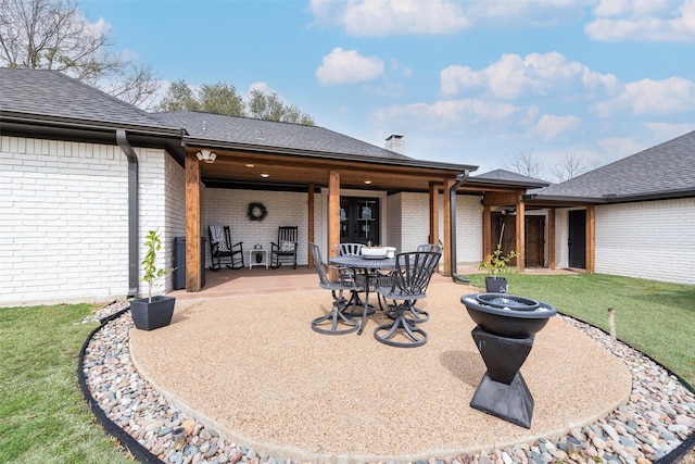 rear view of property featuring a fire pit, brick siding, roof with shingles, a lawn, and a patio area