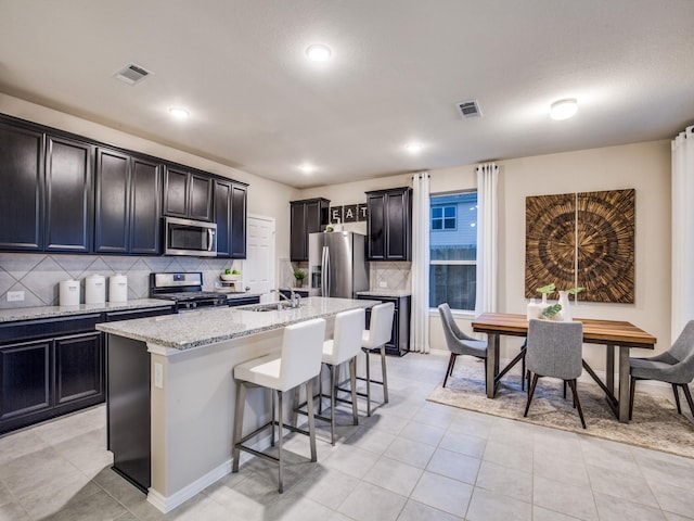 kitchen featuring visible vents, appliances with stainless steel finishes, a kitchen island with sink, light stone countertops, and a kitchen breakfast bar