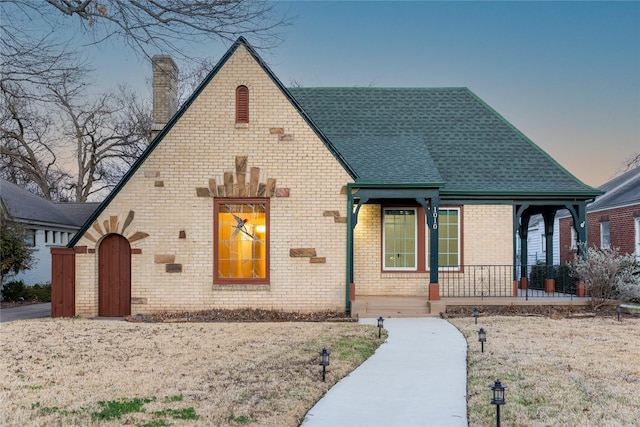 tudor home featuring a shingled roof, covered porch, brick siding, and a chimney