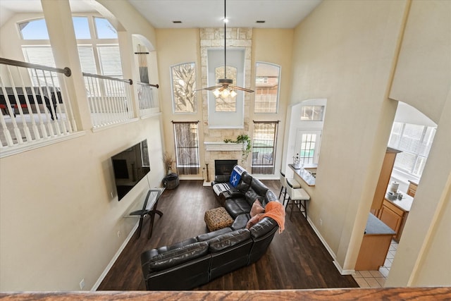 living room featuring ceiling fan, baseboards, a stone fireplace, a high ceiling, and wood finished floors