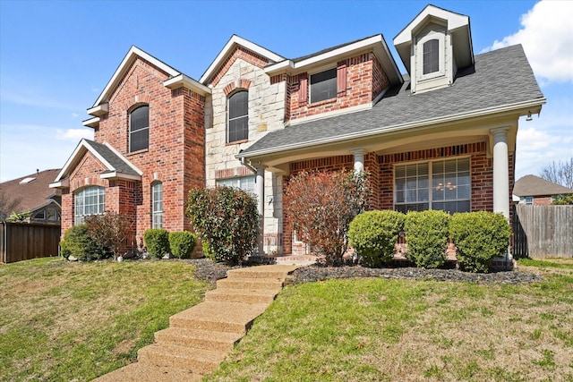 traditional home featuring a front lawn, stone siding, fence, roof with shingles, and brick siding