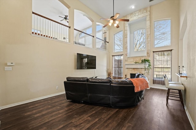 living room featuring a ceiling fan, hardwood / wood-style floors, a stone fireplace, baseboards, and a towering ceiling