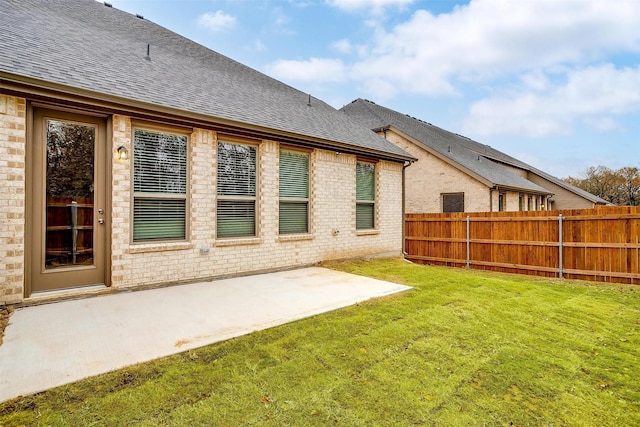 rear view of house featuring a patio, brick siding, fence, roof with shingles, and a lawn