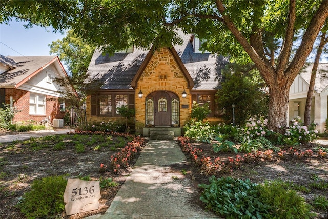 tudor-style house with stone siding, roof with shingles, brick siding, and central AC unit