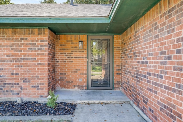 doorway to property featuring brick siding and roof with shingles