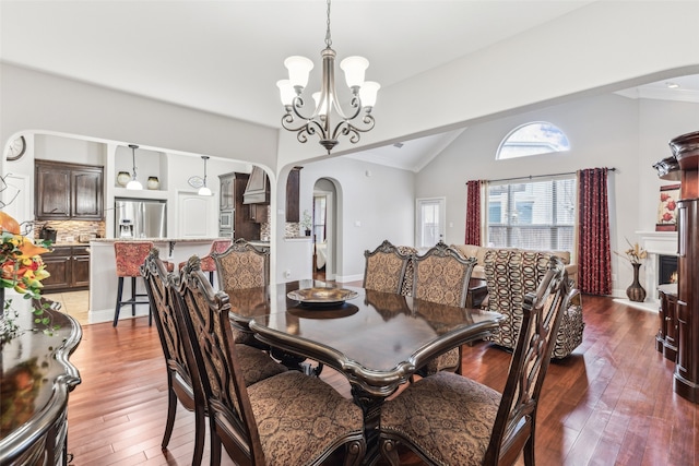 dining room with arched walkways, a lit fireplace, vaulted ceiling, wood-type flooring, and a notable chandelier