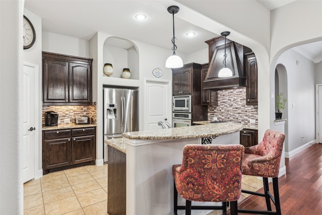 kitchen with dark brown cabinetry, light stone counters, premium range hood, and stainless steel appliances