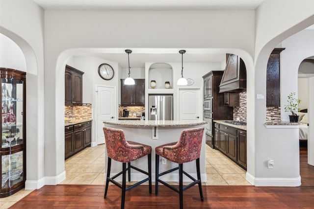 kitchen featuring custom range hood, light stone counters, stainless steel appliances, light tile patterned floors, and dark brown cabinets