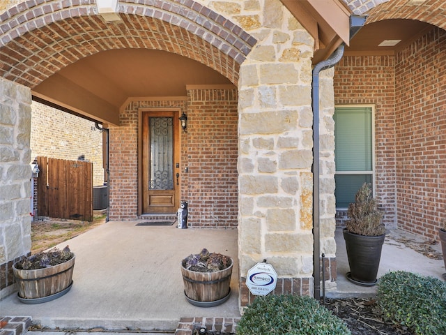 view of exterior entry featuring stone siding, brick siding, fence, and central air condition unit