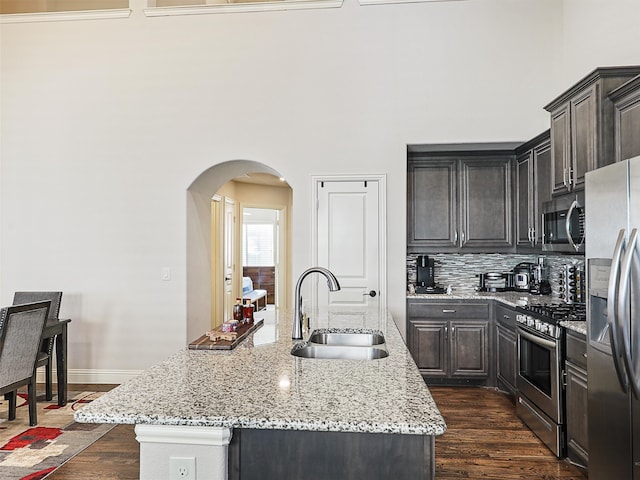 kitchen with dark wood-style floors, arched walkways, stainless steel appliances, a sink, and an island with sink