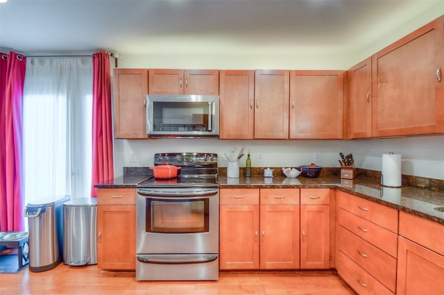 kitchen with light wood finished floors, appliances with stainless steel finishes, brown cabinetry, and dark stone countertops