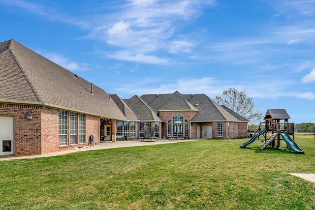 rear view of house featuring roof with shingles, a playground, brick siding, a yard, and a patio area