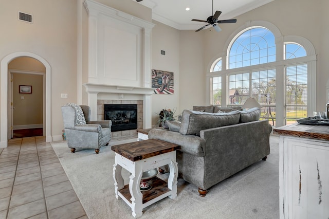 living room with crown molding, a towering ceiling, arched walkways, a tiled fireplace, and a ceiling fan