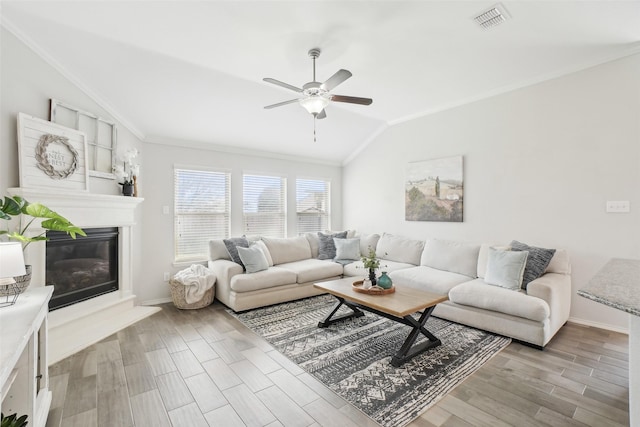 living room featuring visible vents, vaulted ceiling, ornamental molding, light wood finished floors, and a glass covered fireplace