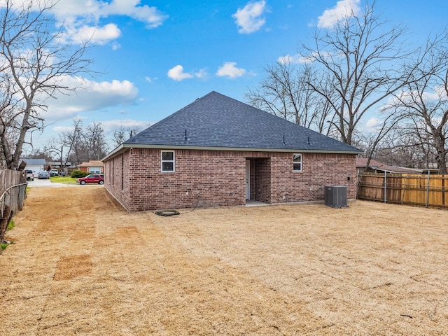 rear view of property with central AC unit, roof with shingles, fence, and brick siding