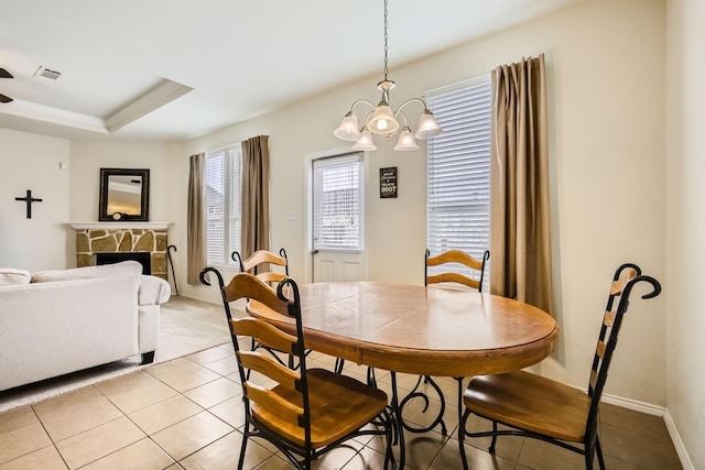 dining area featuring visible vents, a tray ceiling, a stone fireplace, a chandelier, and light tile patterned flooring