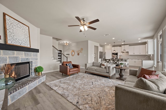 living room featuring baseboards, light wood-style flooring, stairs, a stone fireplace, and recessed lighting