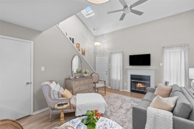 living room with a skylight, light wood-style flooring, a glass covered fireplace, ceiling fan, and high vaulted ceiling