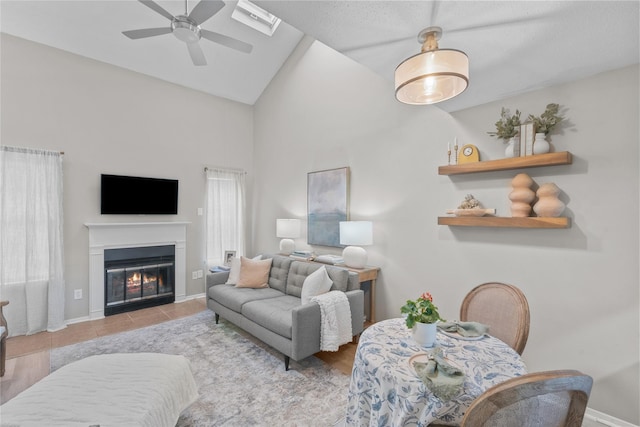 tiled living room with ceiling fan, high vaulted ceiling, a glass covered fireplace, and baseboards