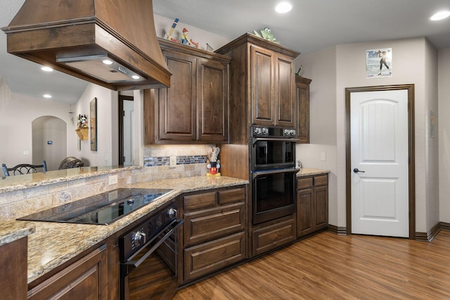 kitchen featuring dark brown cabinetry, tasteful backsplash, custom range hood, wood finished floors, and double oven