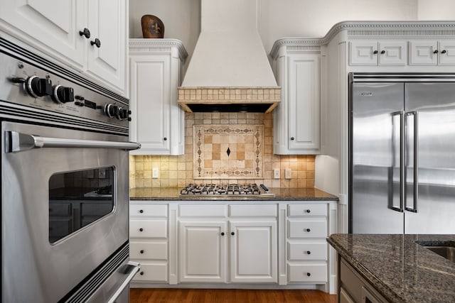 kitchen with dark stone counters, stainless steel appliances, premium range hood, and decorative backsplash