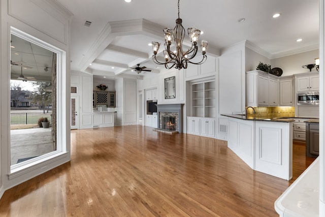 kitchen with a warm lit fireplace, coffered ceiling, a ceiling fan, dark countertops, and stainless steel microwave