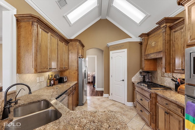 kitchen with light stone counters, arched walkways, stainless steel appliances, brown cabinetry, and a sink