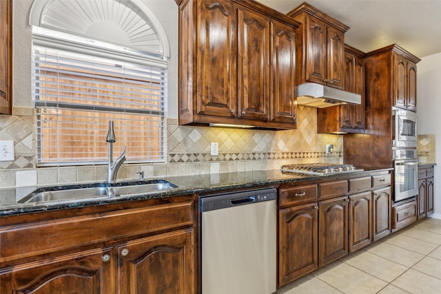 kitchen featuring dark stone countertops, light tile patterned flooring, a sink, under cabinet range hood, and appliances with stainless steel finishes