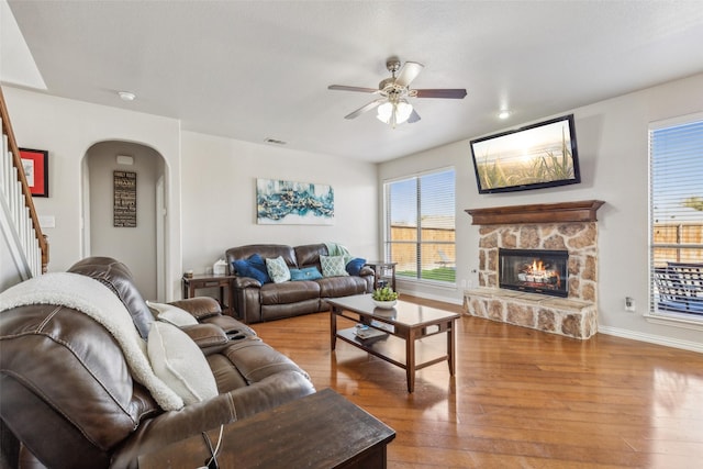 living area with wood finished floors, a ceiling fan, arched walkways, a stone fireplace, and stairs