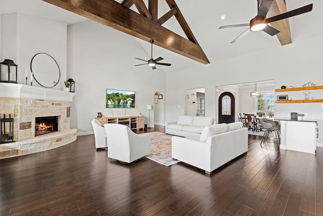 living area featuring ceiling fan, a stone fireplace, dark wood finished floors, and beam ceiling