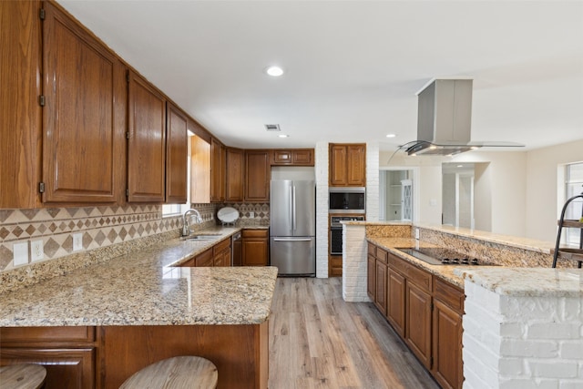 kitchen with decorative backsplash, island range hood, appliances with stainless steel finishes, light stone countertops, and light wood-style flooring