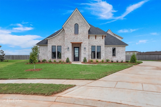 french country inspired facade featuring roof with shingles, brick siding, a front lawn, and fence