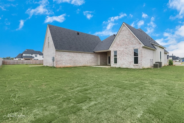 back of property featuring central AC unit, brick siding, fence, roof with shingles, and a lawn