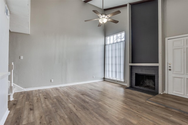 unfurnished living room with baseboards, a ceiling fan, a towering ceiling, wood finished floors, and a brick fireplace