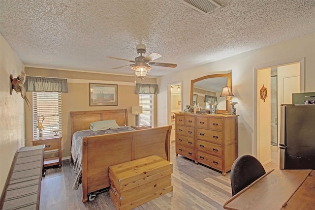 bedroom featuring light wood finished floors, visible vents, freestanding refrigerator, a textured ceiling, and a ceiling fan