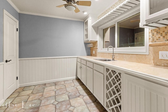 kitchen with a wainscoted wall, crown molding, light countertops, glass insert cabinets, and a sink