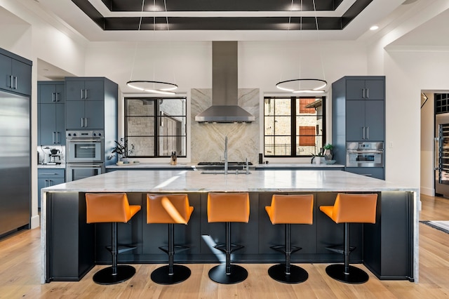 kitchen featuring light wood-type flooring, backsplash, stainless steel appliances, wall chimney range hood, and light stone countertops