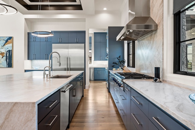 kitchen with blue cabinetry, light stone countertops, wall chimney range hood, and a sink