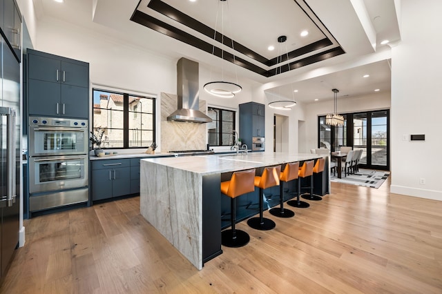 kitchen with a tray ceiling, wall chimney range hood, light wood-type flooring, and stainless steel appliances