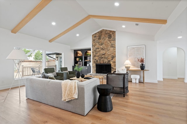 living room featuring arched walkways, beam ceiling, a stone fireplace, and light wood finished floors