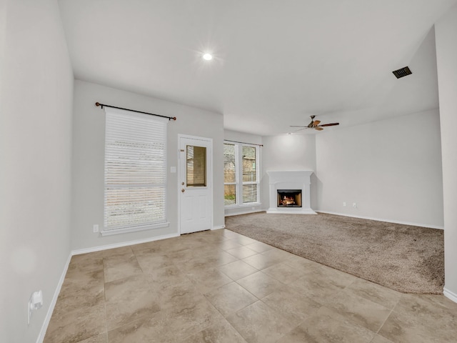 unfurnished living room featuring a warm lit fireplace, carpet floors, visible vents, a ceiling fan, and baseboards