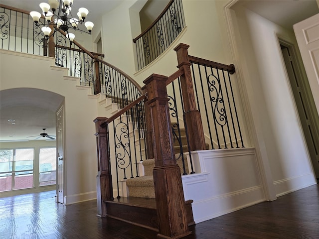 stairs with baseboards, arched walkways, wood finished floors, a high ceiling, and ceiling fan with notable chandelier