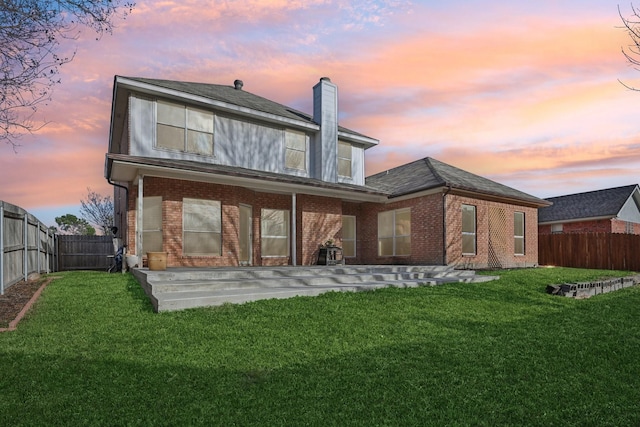 back of house at dusk with brick siding, a lawn, a fenced backyard, and a chimney